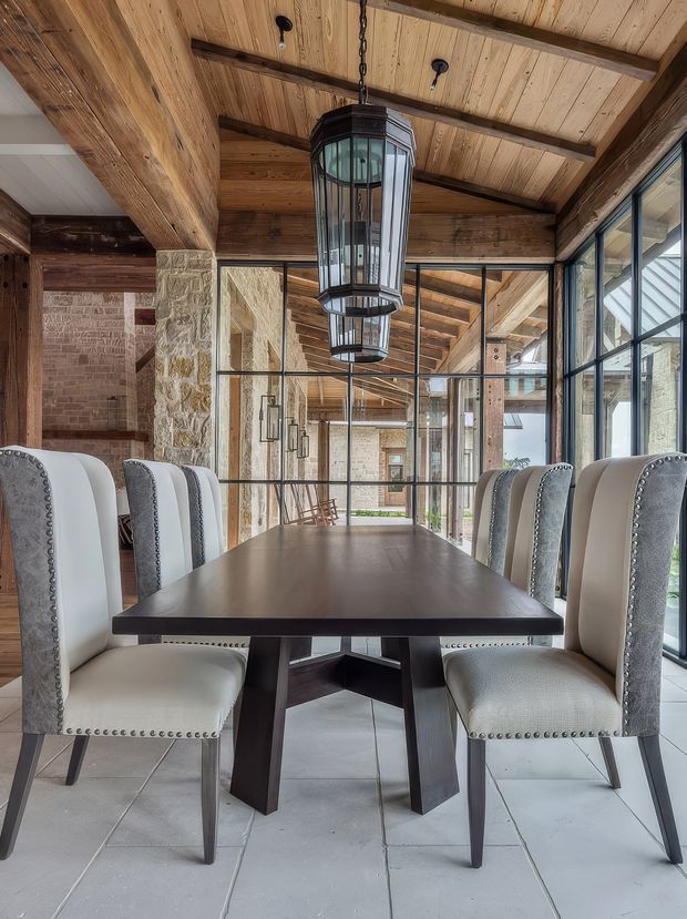 A dining room with a long dark timber dining table surrounded by upholstered nailhead-trimmed chairs. The space features exposed timber ceiling beams and panelling, substantial natural stone and brick elements, black-framed floor-to-ceiling glazing, and large suspended lantern pendants. The glazing reflects and reveals an adjoining timber-and-stone area, reinforcing the open, rustic architectural character.