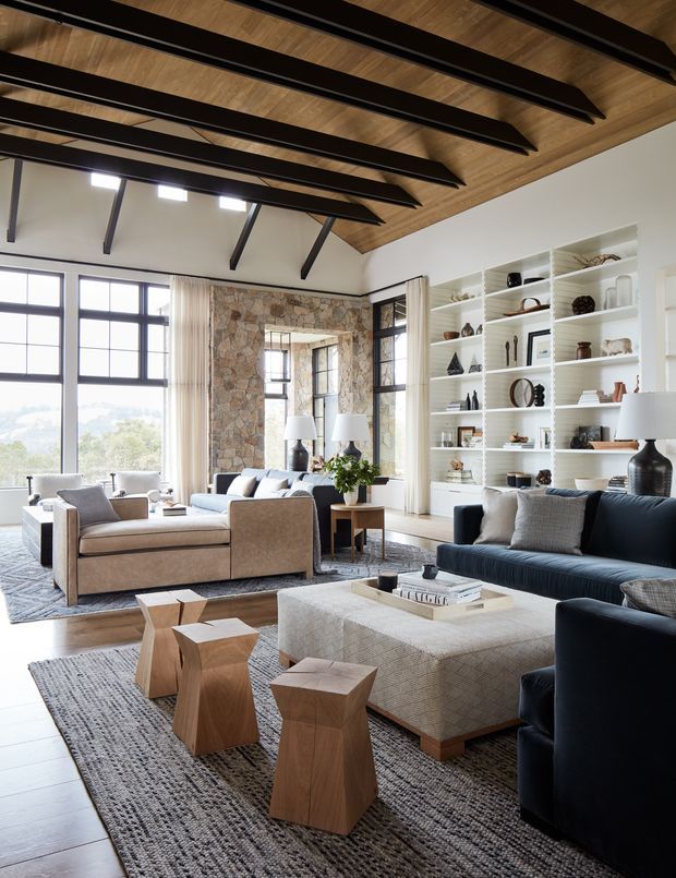 A spacious, light-filled living room with a high sloped ceiling featuring exposed dark timber beams and warm timber lining. Large black-framed windows overlook greenery, while a stone-clad fireplace wall anchors the room. The space includes multiple sofas and armchairs, a large upholstered coffee table, sculptural timber side tables, layered rugs, built-in white display shelving, and neutral contemporary furnishings.