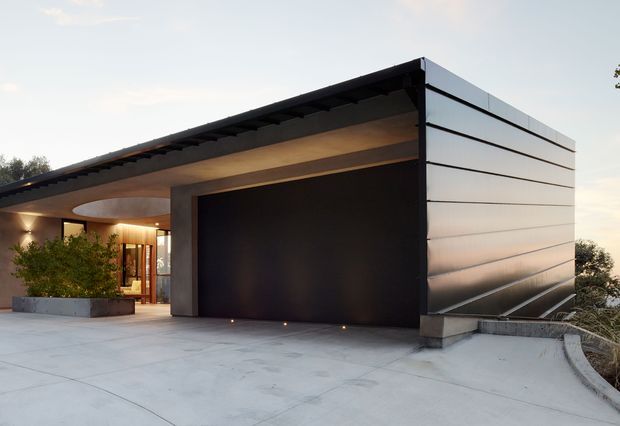 Exterior view of a contemporary single-storey residence with a broad flat roof, dark recessed garage or entry volume, and a light metallic-clad side wall. A curved concrete driveway and low planter define the foreground, while warm interior lighting is visible through glazed entry doors.