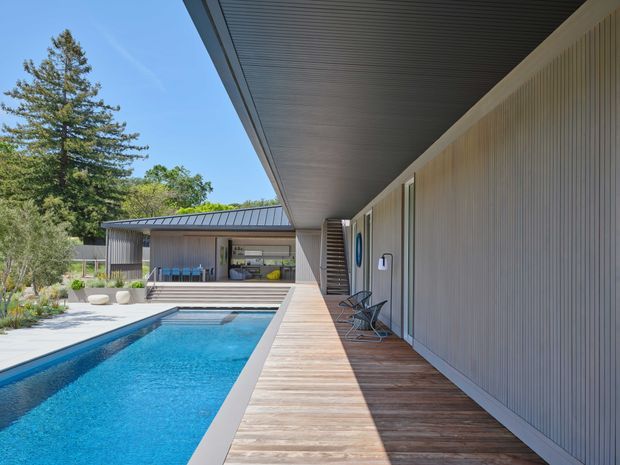 A contemporary residential pool courtyard viewed along a long timber deck beside a swimming pool. The house has a strong horizontal, low-profile form with extensive overhanging roof eaves, vertically ribbed light-coloured wall cladding, dark metal roofing, and large glazed openings. Outdoor dining and lounge areas sit beneath the covered section, with landscaped planting and mature trees beyond.