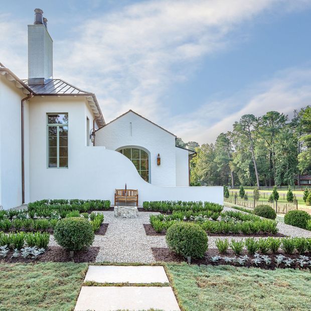 Exterior view of a white-rendered house with a tall metal-clad chimney, steep pitched roof, arched window, and formal landscaped front garden. Gravel paths divide neatly clipped shrubs and low planting beds, leading to a small bench and the house entrance area. A low white garden wall and metal fence frame the property.