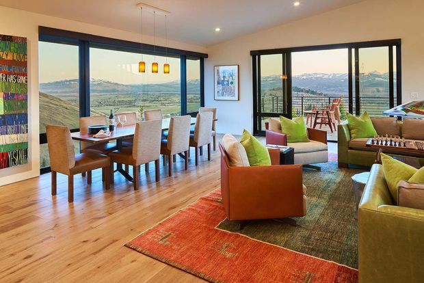 A warm, open-plan living and dining area with expansive black-framed windows overlooking a mountainous rural landscape. The room features timber flooring, a timber dining table with upholstered chairs, a seating area with rust-orange and olive-green upholstered armchairs and sofas, layered area rugs, colourful pendant lights above the dining table, and contemporary artwork. A glazed door opens to an outdoor balcony or terrace with additional seating.