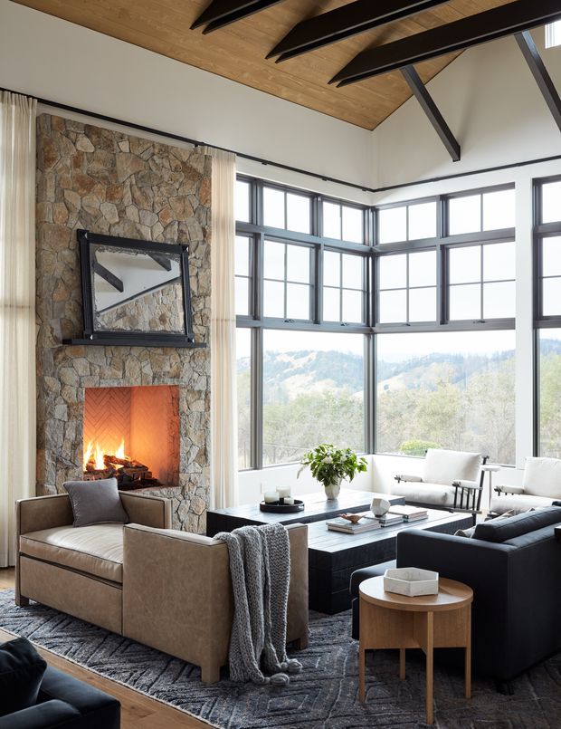 A refined living room with a floor-to-ceiling stone fireplace, lit fire, large black-framed corner windows overlooking a wooded landscape, and a vaulted timber ceiling with exposed dark beams. Neutral upholstered armchairs and sofas surround a dark rectangular coffee table, creating a warm, inviting seating area.