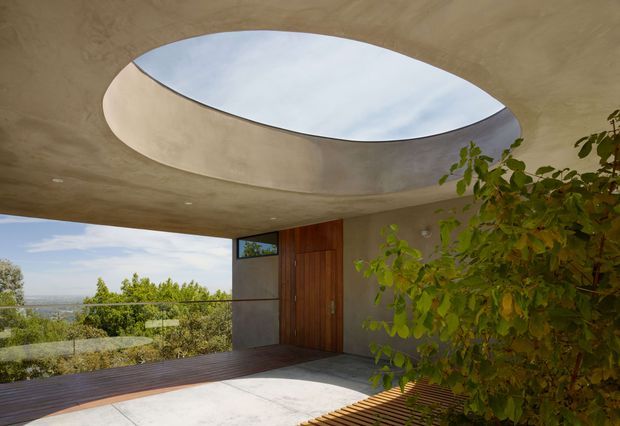 Covered architectural entry or terrace with a sculptural oval skylight set into a smooth curved concrete ceiling. A timber pivot door and dark-framed glazing open toward a leafy elevated landscape, with timber decking and pale concrete flooring creating a restrained indoor-outdoor transition.