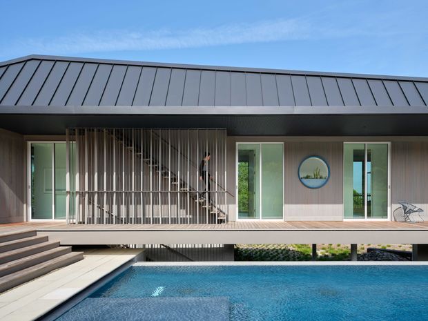 Contemporary single-storey residence viewed from the pool courtyard, featuring a broad dark standing-seam metal roof, timber-toned vertical wall cladding, extensive glazing, and a screened stair enclosure made from closely spaced vertical battens. A raised timber deck and steps overlook a rectangular swimming pool, with landscaped planting visible beneath the elevated structure.