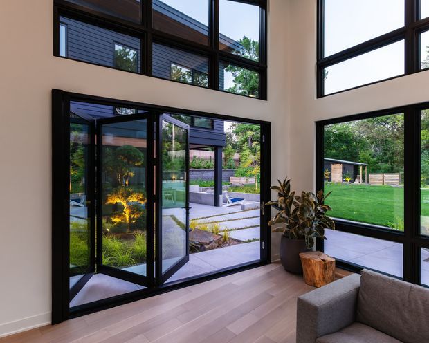 A contemporary double-height living room with expansive black-framed floor-to-ceiling glazing and a large multi-panel glazed door opening onto a landscaped courtyard and lawn. The room has pale timber flooring, light painted walls, a partial grey upholstered sofa, and natural indoor planting with a timber stump side table. The architecture emphasizes strong geometric window grids, abundant daylight, and a seamless indoor-outdoor connection.