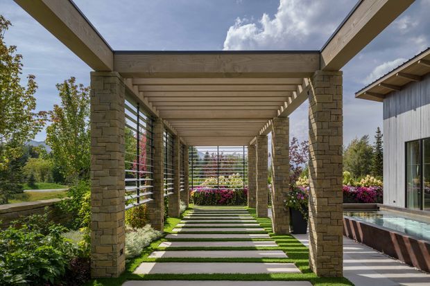 A landscaped outdoor passage beside a contemporary home, framed by a timber pergola with repeated horizontal rafters and substantial stacked-stone columns. Slatted screening encloses the sides and end of the walkway, while alternating concrete stepping slabs and narrow grass strips lead through planted borders. A water feature or pool edge and the home's dark vertical cladding are visible on the right. The scene is bright, refined, and garden-oriented.