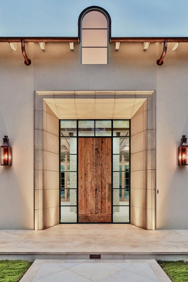 A symmetrical exterior entrance facade with a tall central timber pivot door framed by black metal and glass sidelights, set within a deep pale stone-clad portal. The stucco wall features an arched upper window, copper gutters and downpipes, decorative brackets, and matching wall lanterns. Pale stone paving and a narrow strip of lawn lead to the entry, creating a refined, warm architectural presentation.