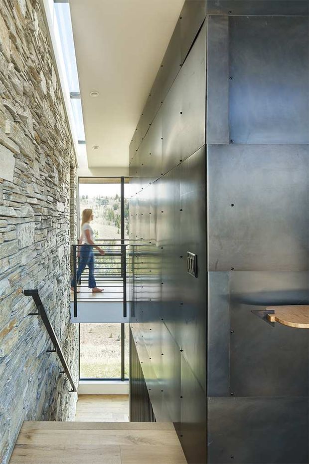 A narrow, vertically oriented interior passage or stair landing with a dramatic material contrast: a rough natural stacked-stone wall on the left and a dark metallic-clad built-in wall or cabinetry on the right. A timber stair or landing occupies the foreground, while a slim black metal balustrade and large floor-to-ceiling glazing lead toward a small intermediate landing with a person beyond. A linear skylight and recessed ceiling lights bring in strong natural light. The design has an industrial, contemporary character with exposed structural and finish materials.