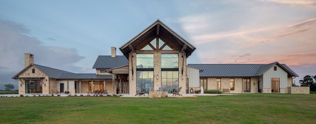 Wide exterior view of a sprawling single-storey residence set behind an expansive lawn. The home features multiple connected wings, steep gabled roof forms with dark metal roofing, substantial stone-clad walls, timber structural detailing, and a prominent central double-height glazed gable. The architecture has a refined rustic lodge or farmhouse character, with a muted natural material palette and sunset sky.