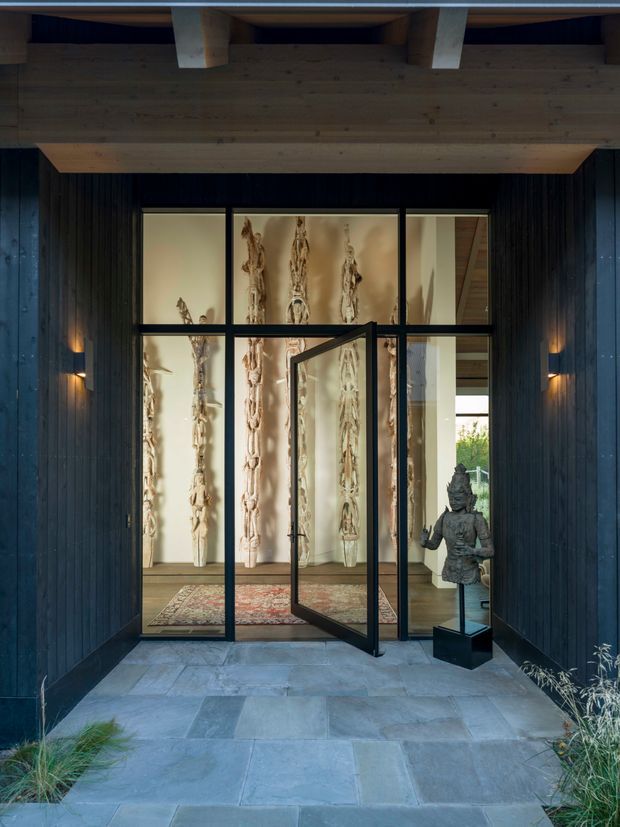 A dramatic covered entry vestibule framed by dark vertical timber cladding, with a broad exposed timber beam overhead and slate-like stone paving underfoot. Black-framed glazed doors open into a refined interior displaying sculptural wall artworks, a rug, timber flooring, and a Buddha statue. Warm integrated wall lighting contrasts with the dark exterior materials, creating a sheltered, gallery-like entrance.