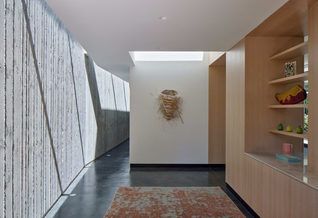 A contemporary residential hallway or entry corridor with a long textured concrete privacy wall and dark polished floor on the left, a skylight above, and full-height warm timber built-in shelving and display cabinetry on the right. A patterned runner, wall sculpture, and curated objects add colour and texture. The space is bright but restrained, with strong linear geometry and a connection to an outdoor passage.