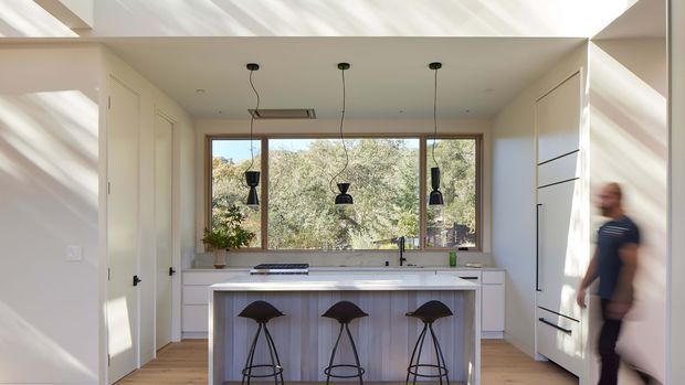 Bright contemporary kitchen with a large central island and three black bar stools, white integrated cabinetry, pale timber flooring, and three black pendant lights. A broad timber-framed window brings in views of leafy vegetation, while strong daylight creates diagonal shadows across the white surfaces.