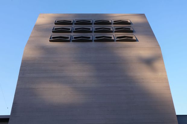 Low-angle exterior view of a tall, monolithic rectangular building facade clad in horizontally lined grey-brown panels. The upper portion features a grid of dark projecting or recessed geometric window openings, while clear blue sky surrounds the structure. The image emphasizes the building envelope and its austere, sculptural composition.