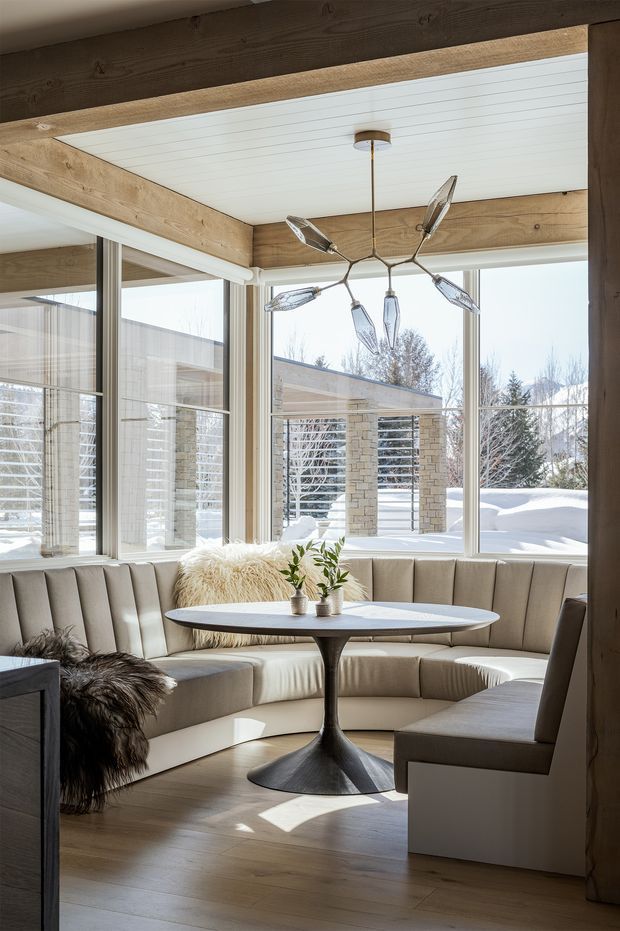 A bright, refined dining nook with a curved upholstered banquette wrapping around a round pedestal table and a separate upholstered chair. Full-height corner glazing overlooks a snowy outdoor setting. Exposed timber beams frame a white plank ceiling, while a sculptural branching pendant light forms the focal point above the table. Pale timber flooring, neutral upholstery, and small potted plants create a warm contemporary interior.