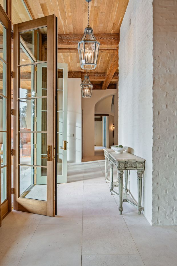 A bright, refined entry hall viewed through an open glazed timber door. The space features a pale stone or porcelain tiled floor, white textured masonry walls, exposed dark timber ceiling beams, warm timber ceiling boards, arched openings, and two hanging lantern pendants. An ornate distressed console table sits against the wall, while glass doors and internal glazing create layered views through the home.