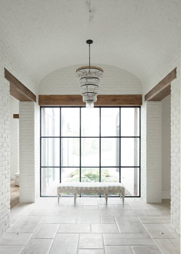 Bright, symmetrical interior space with white-painted brick walls and a vaulted barrel ceiling. A large black-framed industrial-style grid window forms the focal point, with exposed timber beams spanning the room. The pale stone-tile floor, upholstered bench, and tiered crystal chandelier create a refined, airy aesthetic with a blend of industrial and traditional architectural details.