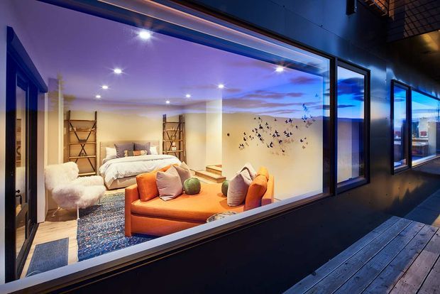 Night-time exterior view through large floor-to-ceiling glazing into a warmly lit bedroom and sitting area. The interior combines a bed, orange sofa, layered rugs, open shelving, and decorative wall art, while the dark exterior facade and timber deck frame the scene. Recessed lighting and blue-purple ambient illumination create a dramatic, boutique-like atmosphere.