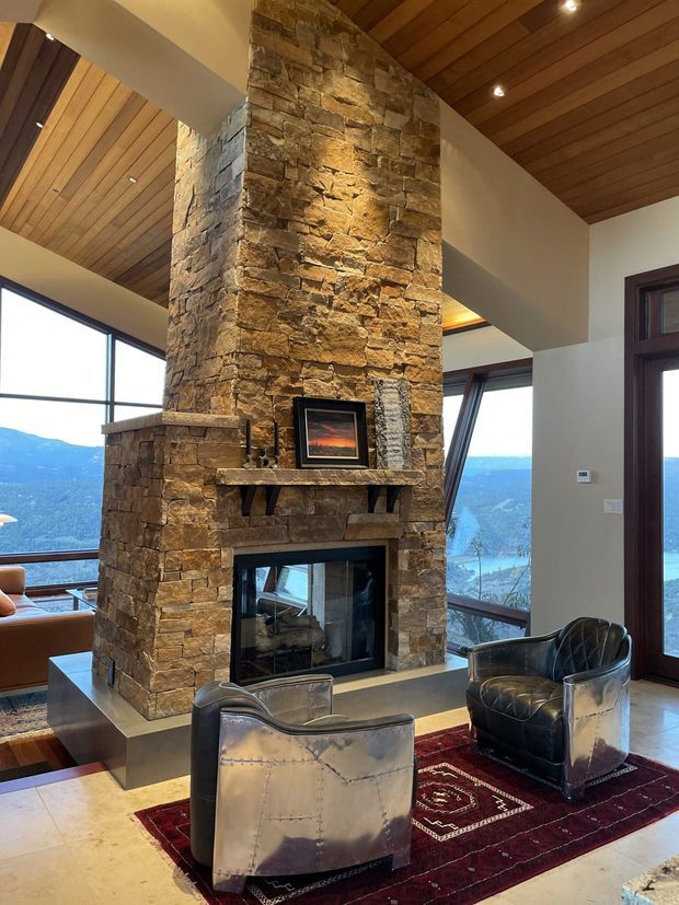 A dramatic mountain-view living room centred on a full-height rough stacked-stone fireplace with a timber mantel and inset firebox. Warm timber-plank ceilings, expansive dark-framed glazing, and a large glazed door open the room to panoramic mountainous and lakeside scenery. Two metallic leather lounge chairs sit on a deep red patterned rug, creating a rustic yet refined lodge-like interior.