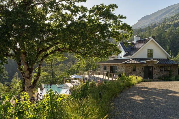 Exterior view of a mountain retreat set among mature trees and landscaped planting. The house has a pitched gabled roof with dark metal roofing, white painted cladding, stone masonry walls, and a broad covered terrace with timber or metal balustrading. A swimming pool is visible below the terrace, while a gravel driveway occupies the foreground. Dramatic wooded mountain terrain forms the backdrop.