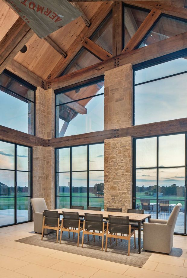 A spacious, light-filled dining area with a long timber dining table and upholstered seating, set beside expansive black-framed glazing. The room features exposed heavy timber post-and-beam construction, a high timber-lined pitched ceiling, and prominent natural stone masonry columns. Views extend to a landscaped rural property outside. The overall character is warm, rustic, and refined, with strong indoor-outdoor connection.