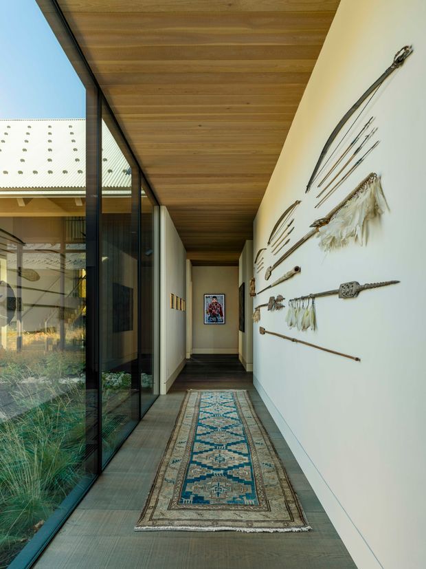 A long, narrow interior hallway with full-height black-framed glazing on one side, opening onto a planted courtyard, and a white display wall on the other decorated with mounted rustic artefacts and textiles. The corridor has a warm timber-lined ceiling, dark flooring, and a patterned runner rug leading toward a framed artwork at the far end. The space is contemporary, linear, and softly lit with a strong indoor-outdoor connection.