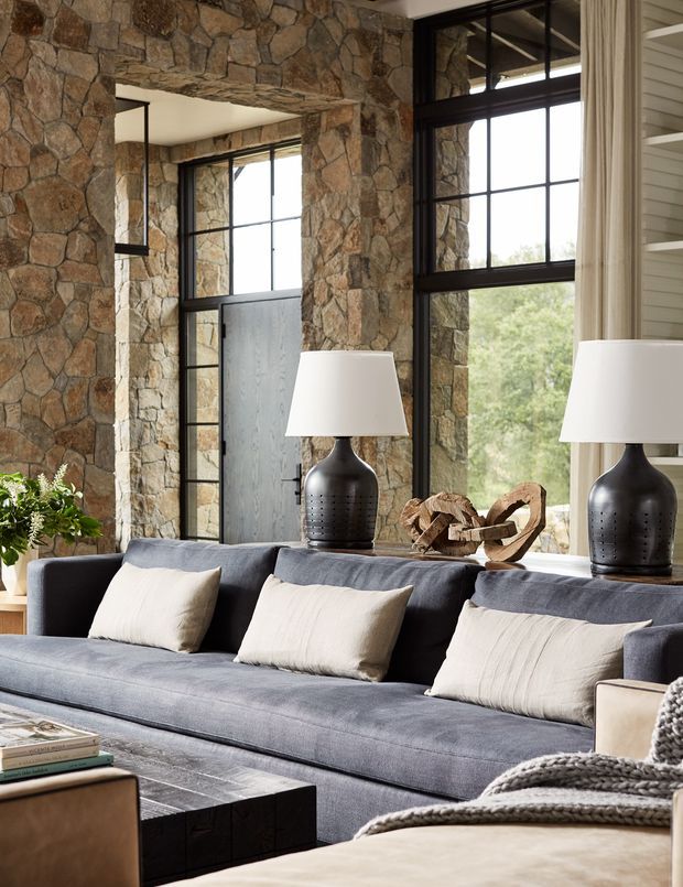 A refined living room with a large slate-blue sectional sofa, cream cushions, dark ceramic lamps, and a low dark coffee table. The room is defined by extensive irregular natural-stone wall cladding, tall black-framed industrial-style windows, and a dark timber or metal entry door. Warm neutral textiles and natural sculptural decor create a sophisticated rustic atmosphere, with greenery visible outside.