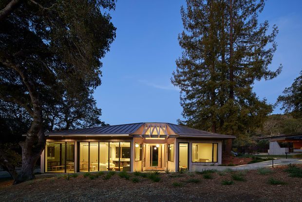 Single-storey residential house viewed from the front at dusk, set among mature trees and landscaped gravel. The low, elongated building has extensive floor-to-ceiling glazing, a dark standing-seam metal roof, warm timber framing and detailing, and a prominent glazed central entry with exposed triangular timber structure. Warm interior lighting is visible through the windows.