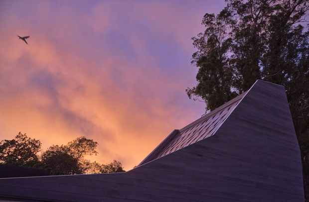 Exterior architectural view at dusk, dominated by a sculptural steeply folded roof form and dark horizontal timber-like cladding. The angular building silhouette is set against a dramatic pink-orange sky with mature trees in the background.