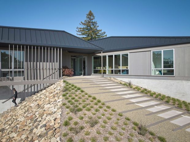 Exterior view of a low, single-storey contemporary building with dark standing-seam metal roofing, grey vertical wall cladding, extensive glazing, and a covered entry. A sloped landscaped approach uses gravel, natural stone retaining and paving slabs, while vertical metal fins screen the glazed portion of the facade.