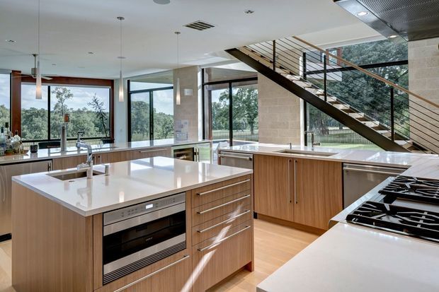 A bright, open-plan contemporary kitchen with two large timber-toned islands and extensive white benchtops. Integrated stainless-steel appliances, a cooktop, sink, pendant lights and extensive cabinetry define the kitchen. Floor-to-ceiling glazing overlooks trees and a garden, while an exposed open-riser staircase with dark metal balustrading crosses the background.