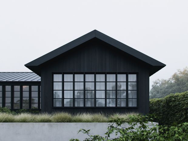 Exterior view of a dark, single-storey gabled building with vertical black cladding and a broad grid of black-framed windows. A lower glazed wing extends to the left, while layered grasses, hedging, and a pale rendered retaining wall form the foreground landscaping. The image has a restrained, contemporary rural-inspired appearance under overcast light.
