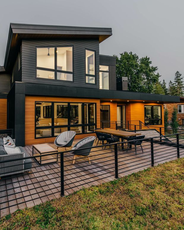 A modern two-storey residential home viewed from an elevated outdoor deck. The facade combines dark horizontal cladding and trim with warm timber-look wall panels, extensive black-framed glazing, and a shallow projecting roof. The spacious deck has black metal railings, timber decking, outdoor lounge chairs, a dining table, and built-in seating, with planted landscape in the foreground.