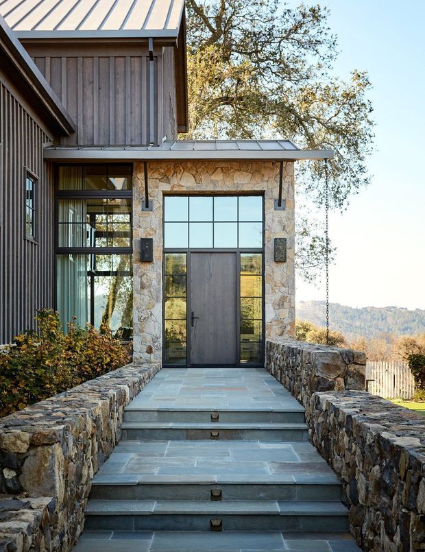 Exterior approach to a contemporary home entry, featuring a stone-clad doorway with a dark timber door and large gridded glazing, flanked by low rough-stone retaining walls and broad slate-like steps. The facade combines vertical timber cladding, standing-seam metal roof planes, extensive glazing, and natural stone. A wooded, hilly landscape is visible beyond.