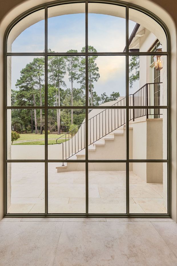 Interior view through a large arched, multi-pane steel-framed glazed door or window. Beyond the glazing is a pale stone-paved terrace with an exterior staircase, dark metal balustrades, cream rendered walls, and landscaped trees. The image emphasizes the dramatic arched opening, generous glazing, and connection between interior and exterior.