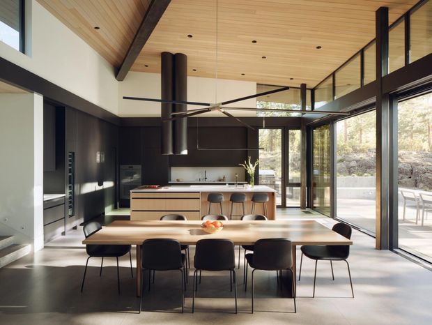 A spacious, contemporary open-plan kitchen and dining area with extensive floor-to-ceiling glazing. The room features a long timber dining table with dark chairs, a central kitchen island with seating, dark integrated cabinetry, a large sculptural ceiling fan, black structural beams and frames, and a warm timber-lined sloping ceiling. Outdoor landscaping is visible through the glazed doors.