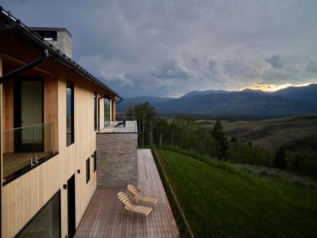 Exterior view of a contemporary multi-level timber-clad house on a sloping site, with a long timber deck and outdoor loungers overlooking a broad mountain valley. The building has a dark pitched roof, large windows, a stone-clad projecting wall, and a dramatic cloudy mountain landscape beyond.