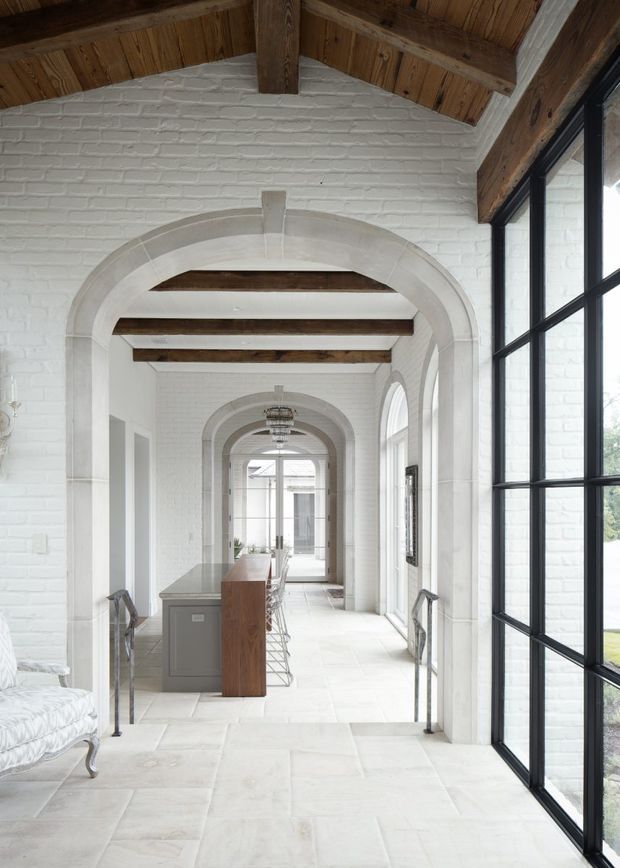 A bright, elegant interior corridor or open-plan circulation space with repeated white-painted brick arches, pale stone flooring, exposed dark timber beams, and large black-framed windows. A compact kitchen or bar area with timber cabinetry and stools is visible along the left side, while the long arched vista leads toward glazed doors at the far end. The room has a refined, airy, heritage-inspired character with strong architectural rhythm.