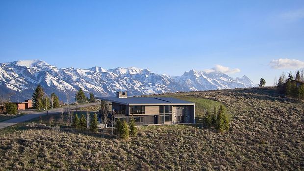 A modern single-storey house set on an open sloped hillside, with extensive glazing, a low-profile dark roof, and a mountain range dominating the background. The surrounding landscape is sparse and natural, with a driveway, scattered trees, and rugged alpine terrain.