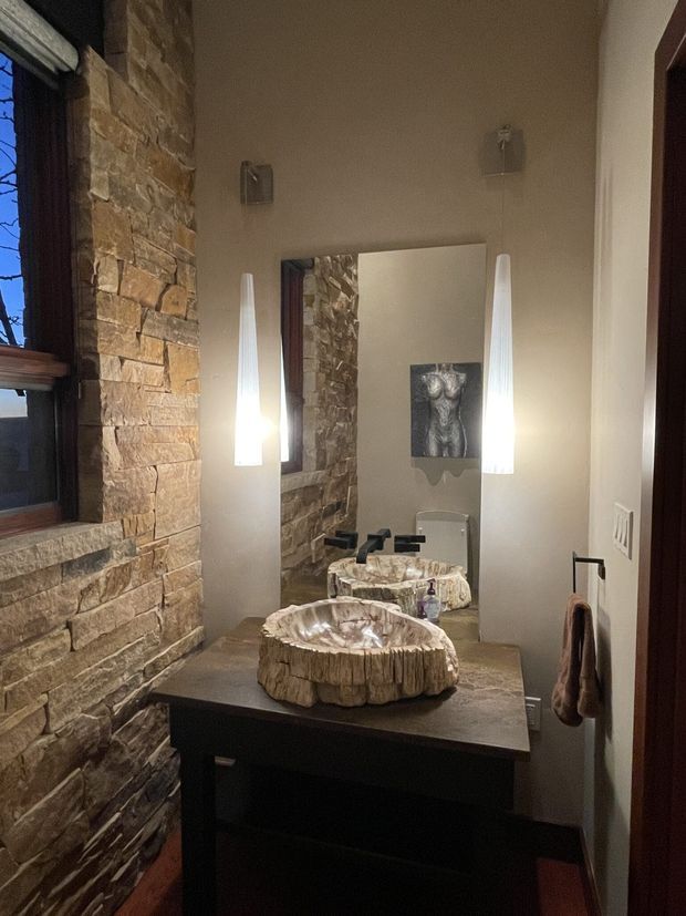 A compact, moody bathroom with a dark timber vanity, stone-clad wall, large rectangular mirror, twin sculptural vessel basins, and black wall-mounted taps. Vertical illuminated pendants flank the mirror, while a small window and matching stone detailing add a rustic, natural character.