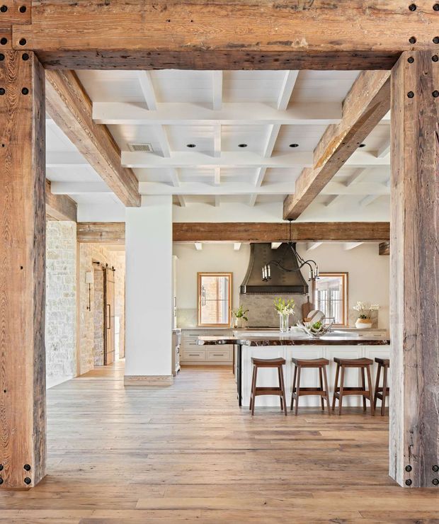 Bright open-plan kitchen viewed through a substantial timber-framed opening, featuring exposed rustic wood beams, white coffered ceiling panels, pale timber flooring, light cabinetry, a stone island benchtop, and dark wood bar stools. A large dark metal rangehood forms a focal point, while stone walling and warm neutral finishes reinforce the farmhouse character.