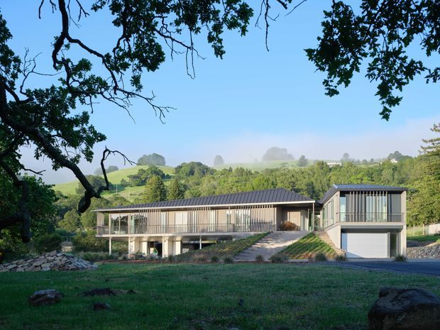 A contemporary single-storey house set in a landscaped rural, rolling-hills setting. The home has a long low profile, dark pitched metal roof, extensive floor-to-ceiling glazing with vertical screening, elevated living areas, a covered deck or balcony, and an integrated garage at the right. Stone retaining walls and a broad lawn occupy the foreground, with mature trees framing the view.