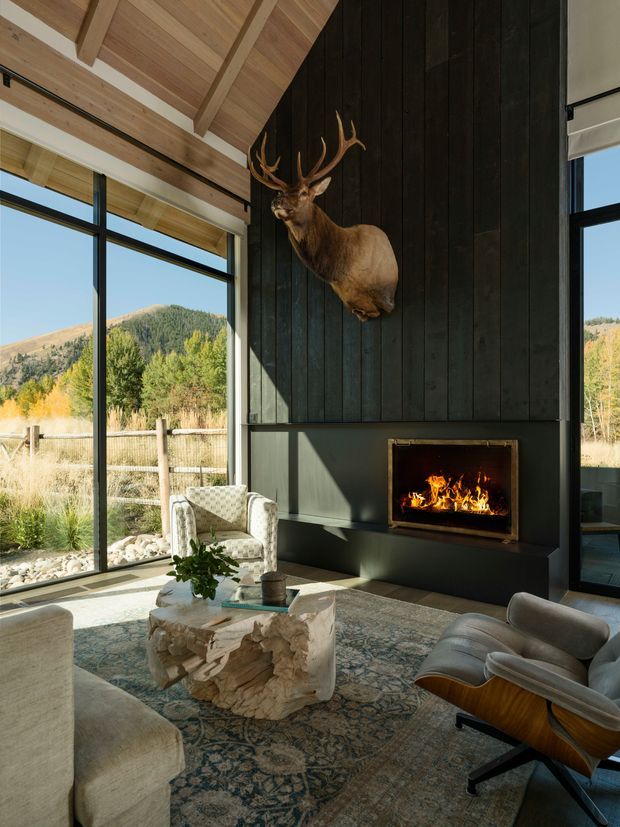A spacious, light-filled living room with floor-to-ceiling black-framed glazing overlooking a mountainous landscape. The room features a dramatic black vertical timber feature wall with a built-in fireplace, a vaulted timber-lined ceiling with exposed beams, neutral upholstered seating, a sculptural natural wood coffee table, and a mounted deer head. The overall aesthetic is warm, rustic, and contemporary, with strong indoor-outdoor connection.