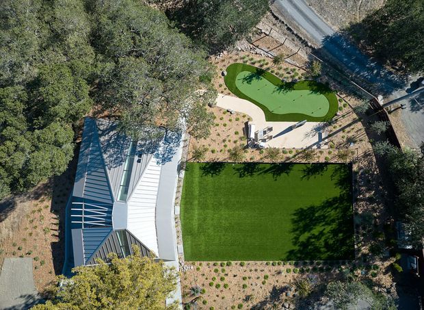 Aerial view of a landscaped residential property surrounded by mature trees. A light-coloured house with a pitched metal roof sits beside a large rectangular lawn, drought-tolerant planting beds, paved outdoor seating areas, and a small curved green swimming pool. A narrow road runs along the upper edge of the site.