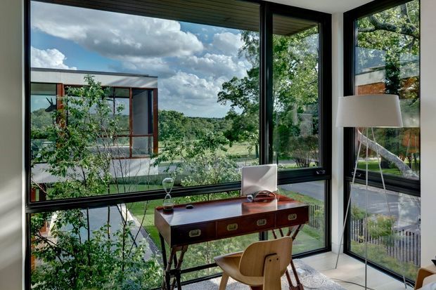 A contemporary home office or study positioned beside expansive corner glazing, with a compact mid-century-inspired wooden desk, wooden chair, desk lamp, and sculptural decor. Black-framed floor-to-ceiling windows overlook a leafy landscaped site and reveal another modern pavilion-like structure outside. The room has light neutral walls and flooring, strong indoor-outdoor connection, and abundant natural light.