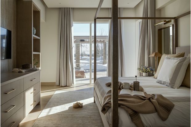 A bright, refined bedroom with a timber four-poster bed, layered neutral bedding, built-in timber-toned cabinetry and shelving, a wall-mounted television, and full-height curtains framing glazed doors to an outdoor terrace. The room features warm natural light, pale walls, a large light-toned rug, and understated contemporary furnishings.