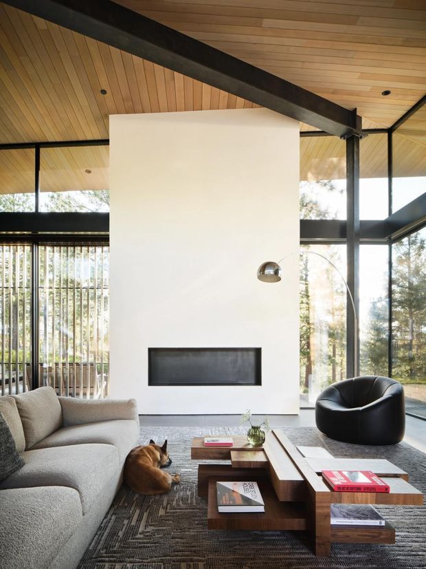 A light-filled contemporary living room with extensive floor-to-ceiling glazing, a white fireplace wall with a slim black firebox, exposed dark steel beams, and a warm timber-lined ceiling. The room contains a large neutral sectional sofa, a sculptural black lounge chair, layered timber coffee tables, and a patterned dark rug. Vertical exterior screening and trees are visible beyond the glazing.