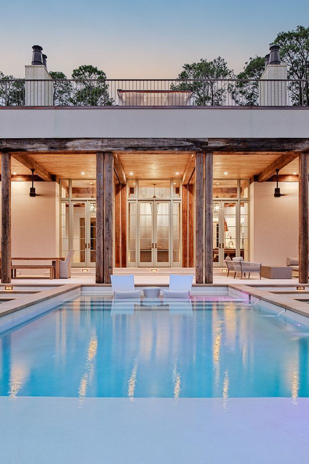 A twilight view across a rectangular swimming pool toward a refined covered outdoor living space. The pavilion features substantial weathered timber posts and beams, warm timber soffit ceilings with recessed lighting, pale rendered walls, and large multi-pane glazed doors. A raised parapet and rooftop railing sit above the pavilion, with masonry chimney-like structures at both sides. Poolside loungers and understated outdoor seating are visible beneath the covered area, creating a calm, luxurious resort-like setting.