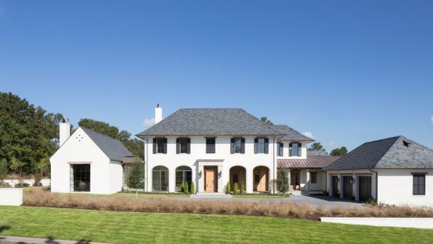Exterior view of a large traditional two-storey residence with pale rendered walls, dark shutters, arched ground-floor openings, a central entry, and multiple grey hipped and pitched roof forms. The house includes attached side wings and a broad manicured lawn in front under a clear blue sky.