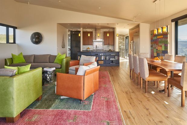 Open-plan living, dining and kitchen interior with a warm contemporary design. The room features timber flooring, a dark kitchen island and cabinetry, stone accents, a large dining table, upholstered dining chairs, grey and vivid green sofas, orange leather armchairs, and colourful patterned rugs. Large windows provide expansive exterior views, while recessed ceiling lights and pendant lighting illuminate the space.